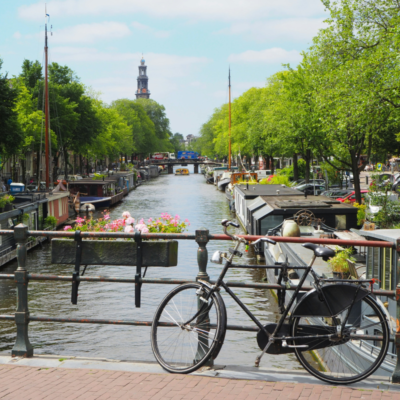 Amsterdam-canal A black bicycle parked on a bridge overlooking an Amsterdam canal filled with houseboats and lined with trees.