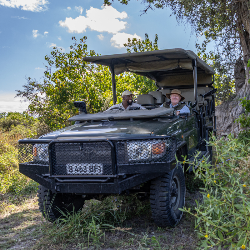 An open-top dark green safari vehicle with a driver and a passenger driving through a grassy area.