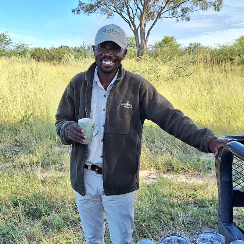 A smiling safari guide wearing a fleece jacket and a cap, standing in a grassy field holding a mug.