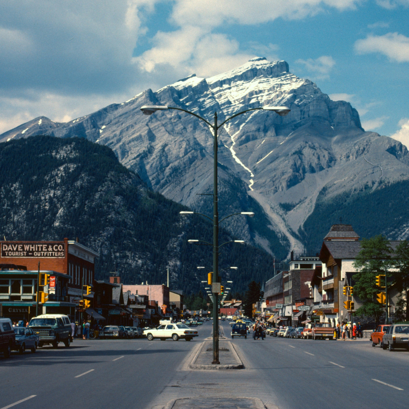 Looking down a wide town street lined with vintage storefronts, cars, and lampposts toward a towering, snow-dusted mountain.