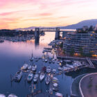 A marina filled with yachts and boats at dusk, with modern apartment buildings and a bridge under a pink and purple sunset sky.