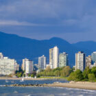 A sandy beach and blue ocean waters in the foreground with the Vancouver city skyline and large mountains behind it.