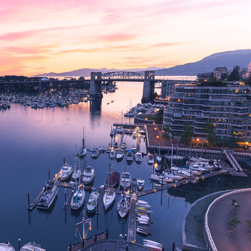 A marina filled with yachts and boats at dusk, with modern apartment buildings and a bridge under a pink and purple sunset sky.