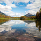 A perfectly still lake reflecting the blue sky, clouds, and autumn forests stretching toward distant mountains.