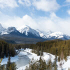 A winding snow-lined river curves through a pine forest toward snow-capped mountains under a cloudy sky.