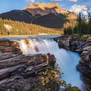 Waterfall cascading through rocky cliffs into a turquoise river, with a dense pine forest and a massive sunlit mountain in the background.