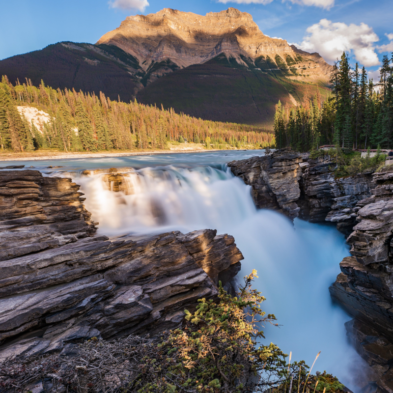 Canada-waterfall Waterfall cascading through rocky cliffs into a turquoise river, with a dense pine forest and a massive sunlit mountain in the background.