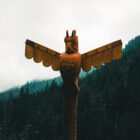 A close-up of a carved wooden totem pole shaped like an eagle with outstretched wings against a foggy forest background.