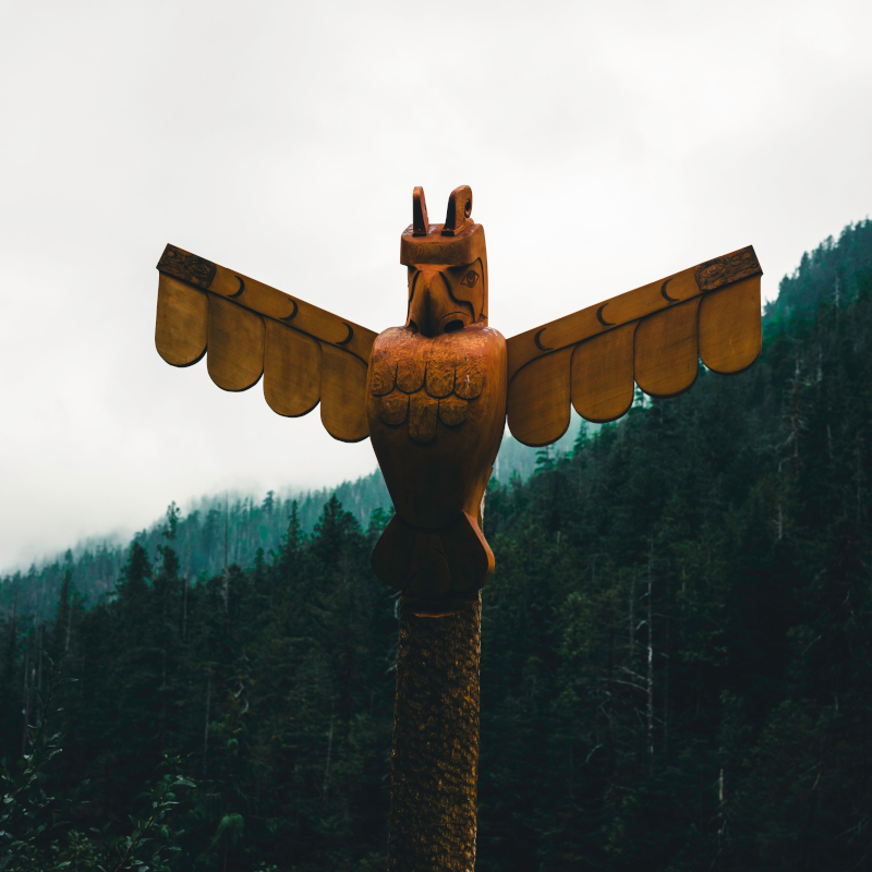 A close-up of a carved wooden totem pole shaped like an eagle with outstretched wings against a foggy forest background.