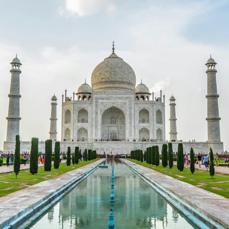 India-taj-mahal A symmetrical, front-facing view of the white marble Taj Mahal with its reflection visible in the long garden pool in front of it.