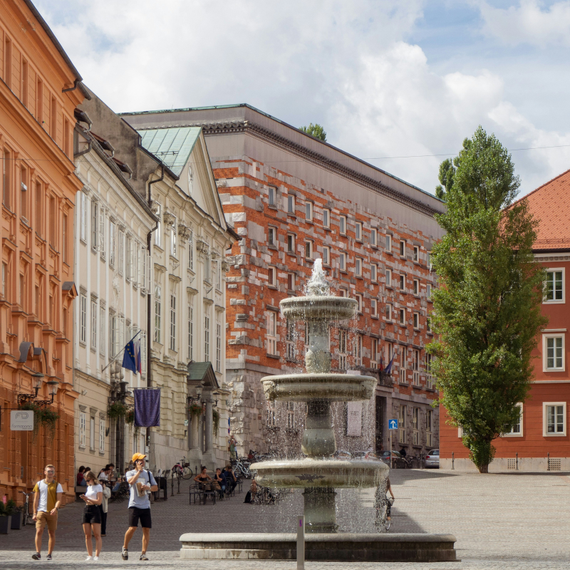 Ljubljana-Slovenia A tiered stone water fountain in a European city square, surrounded by colourful historic buildings and pedestrians.