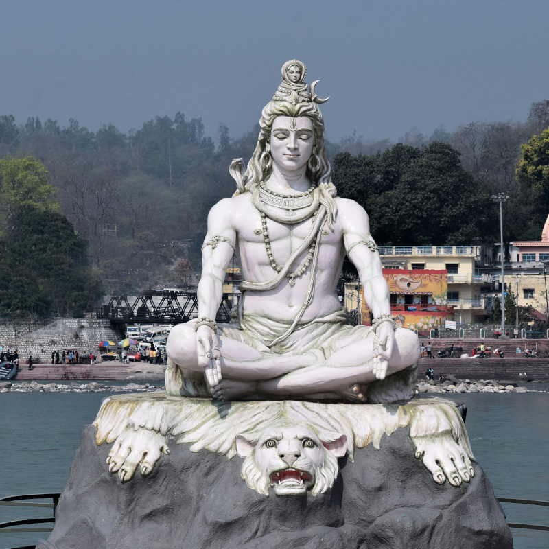 Lord-Shiva-statue Large white stone statue of Lord Shiva sitting in a meditative lotus position beside a body of water.