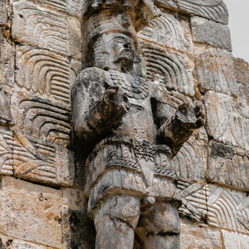 Ancient stone carving of a warrior or deity built into a detailed relief wall at a Mayan archaeological site.