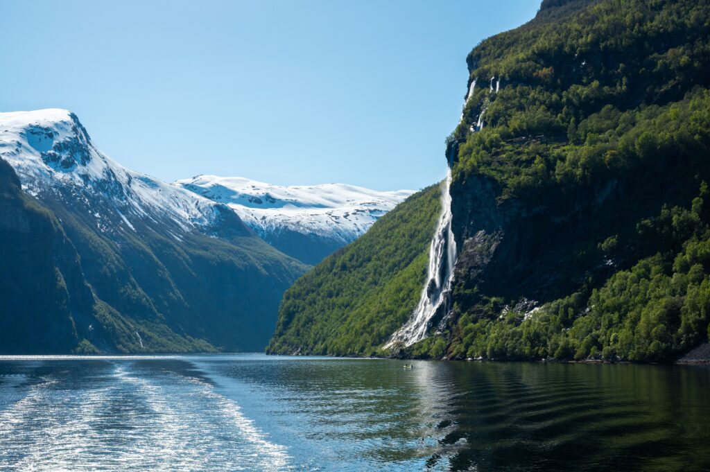 Norway-Fjords A dramatic waterfall cascading down a sheer, green cliff into a deep Norwegian fjord, with snow-capped mountains in the distance.