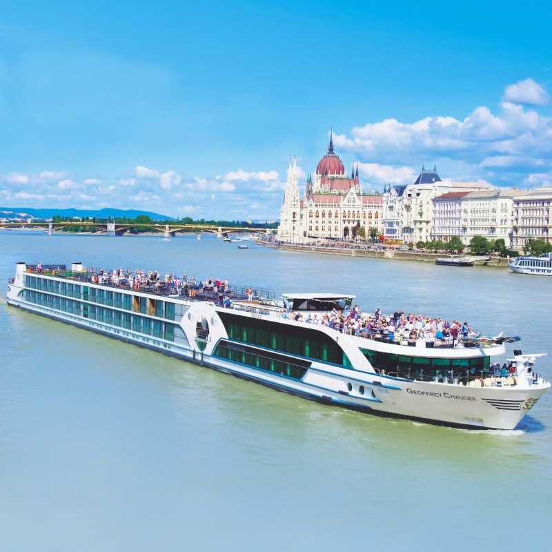 River-cruise-Riviera A large, multi-deck river cruise ship carrying passengers past a grand European palace.
