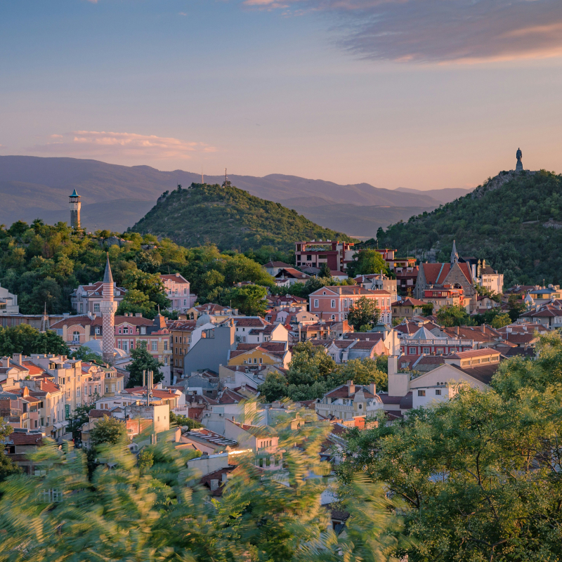 Scenic-city-view A hillside European city view at sunset, showing clustered buildings with orange roofs, a minaret, and lush green hills.