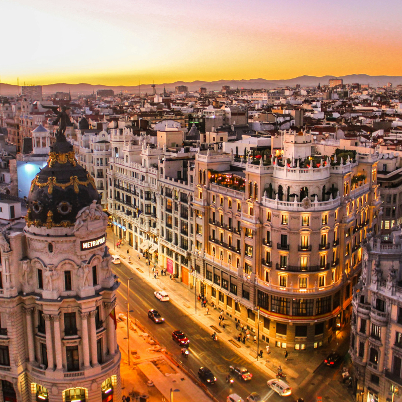 A high-angle view of the iconic Metropolis Building and Gran Vía street in Madrid, illuminated at sunset with city traffic below.