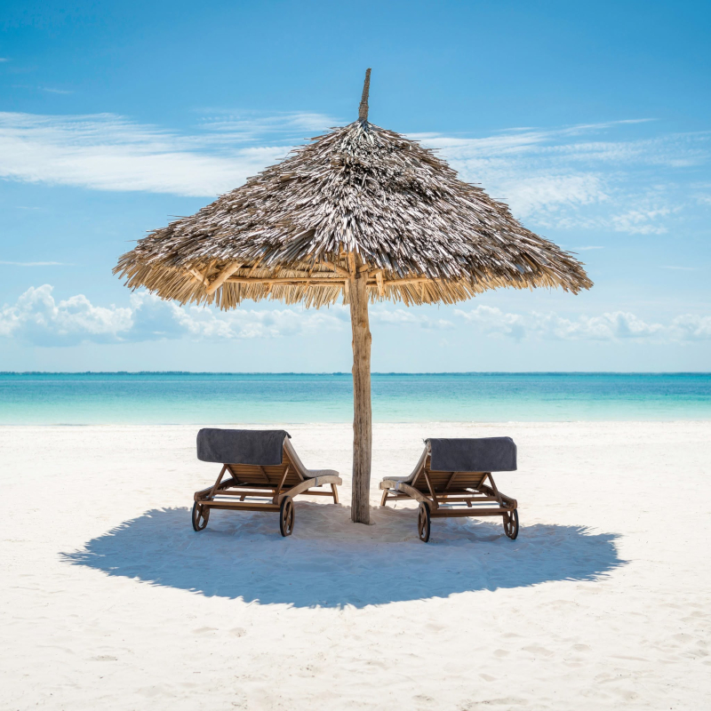 Two wooden lounge chairs with grey towels sit in the shade of a large, thatched umbrella on a white sand beach facing a turquoise ocean.