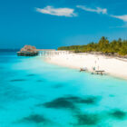 High-angle view of a tropical white-sand beach in Zanzibar, featuring vibrant turquoise water, a wooden pier structure, and small boats