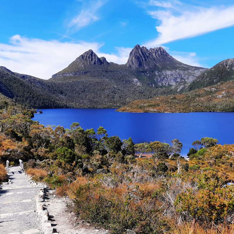 A stone pathway leading toward a vibrant blue lake with jagged mountain peaks in the background.