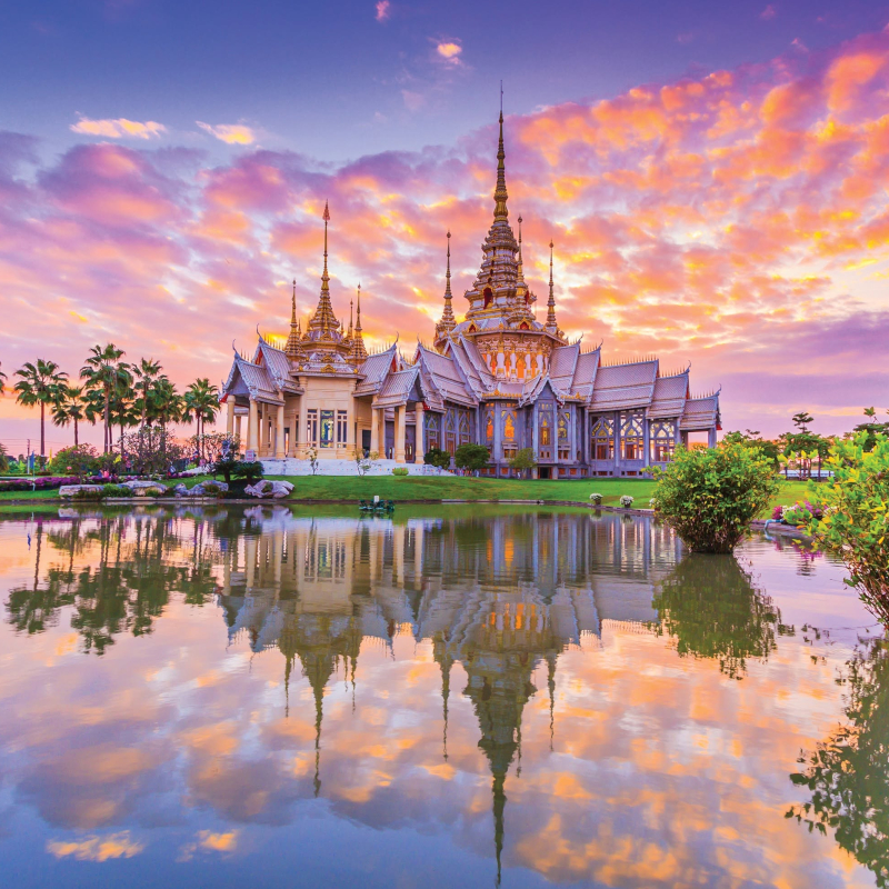 A grand, ornate Thai temple with golden spires is perfectly reflected in a calm pond during a vibrant pink and orange sunset.