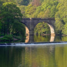 A stone arch bridge spans across a calm river reflecting green trees, with a small weir creating rushing white water in the foreground.