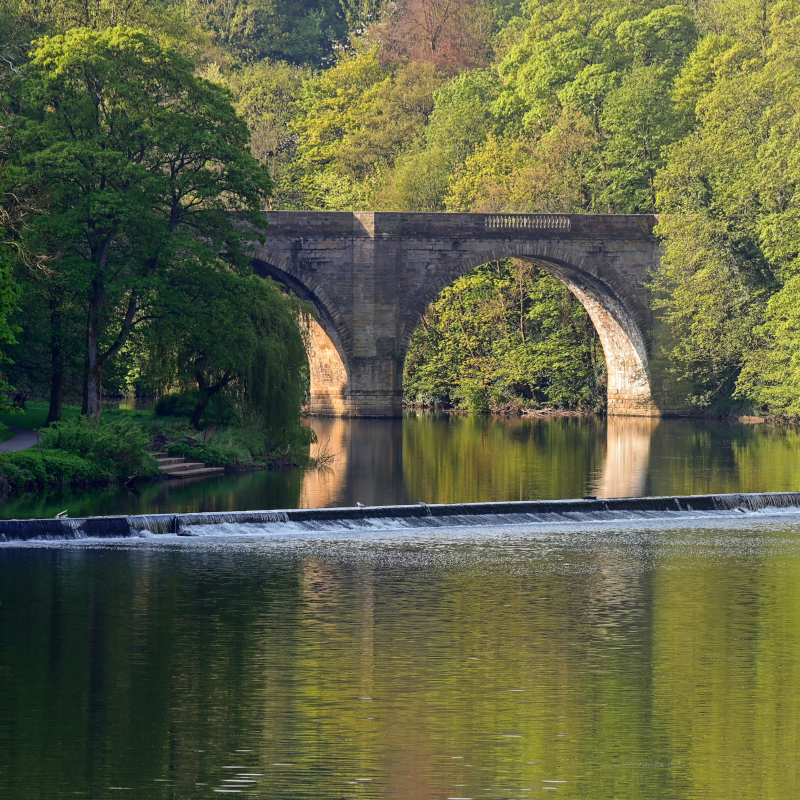 A stone arch bridge spans across a calm river reflecting green trees, with a small weir creating rushing white water in the foreground.