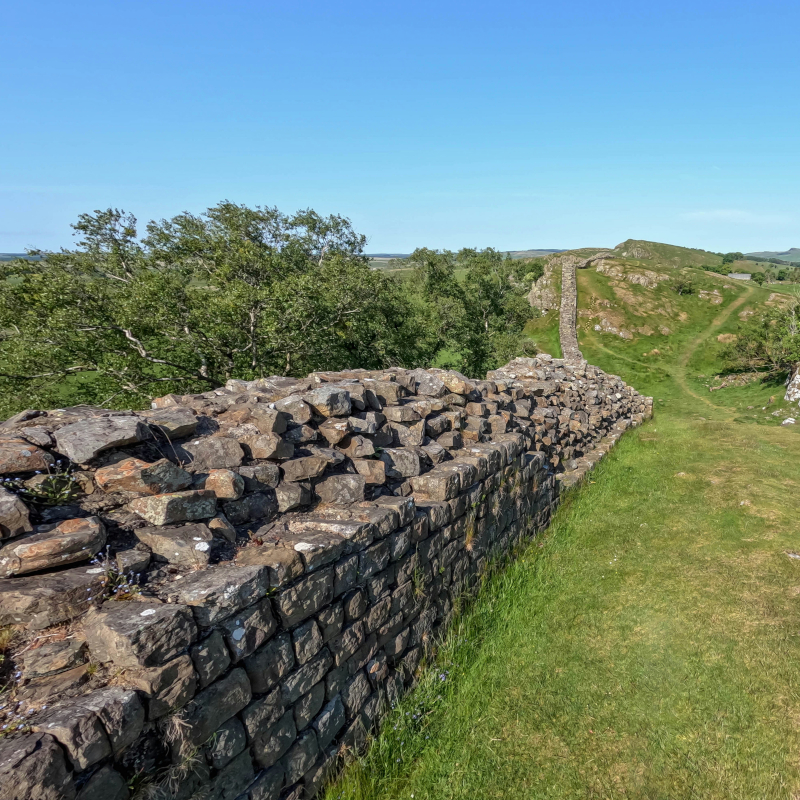 An ancient stone wall winds its way over rolling green grass hills under a clear blue sky.
