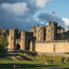A large stone medieval castle with a Union Jack flag flying from a tower, featuring a stone wall, green lawns, and visitors under a cloudy sky.