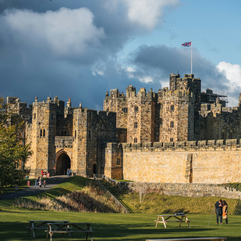 A large stone medieval castle with a Union Jack flag flying from a tower, featuring a stone wall, green lawns, and visitors under a cloudy sky.