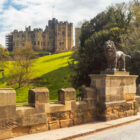 A view looking up a green grassy hill toward a massive stone castle, with a stone wall and a prominent lion statue on a pedestal in the foreground.