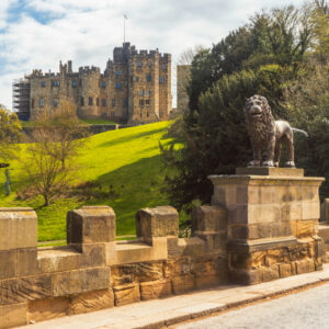 A view looking up a green grassy hill toward a massive stone castle, with a stone wall and a prominent lion statue on a pedestal in the foreground.