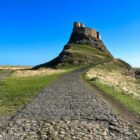 A cobblestone path curves upward through a green field toward a stone castle situated on top of a steep rocky hill under a clear blue sky.
