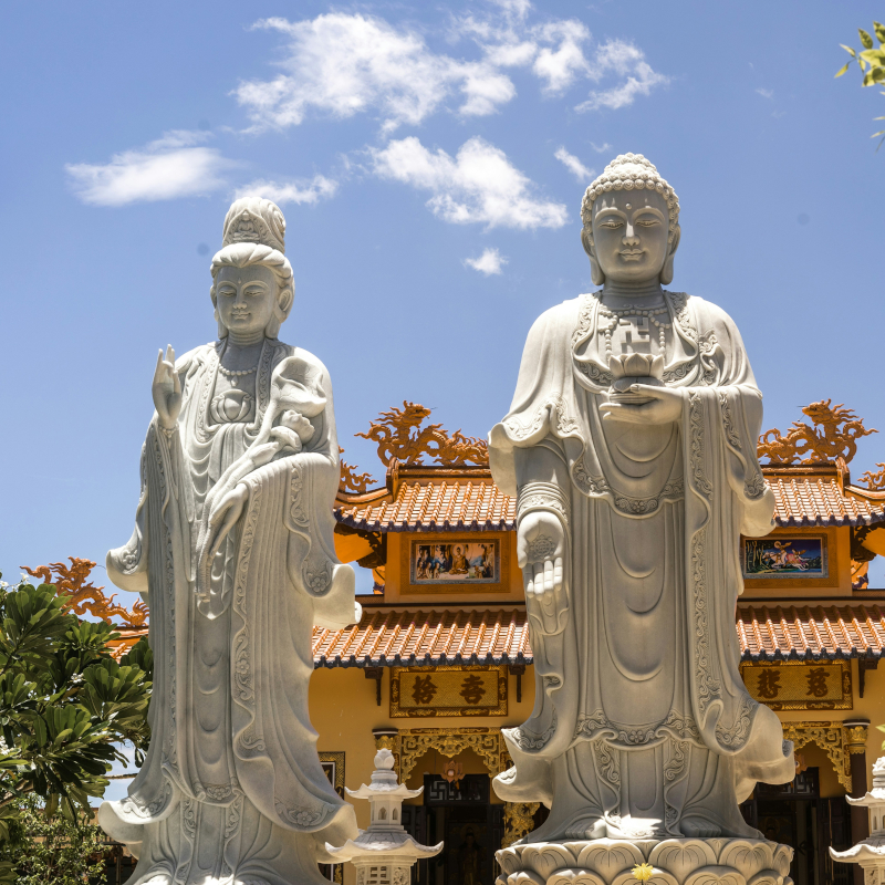 White-marble-Buddhist-statues (1) Two large, detailed white marble Buddhist statues stand in front of a traditional yellow temple with an ornate orange tiled roof against a blue sky.