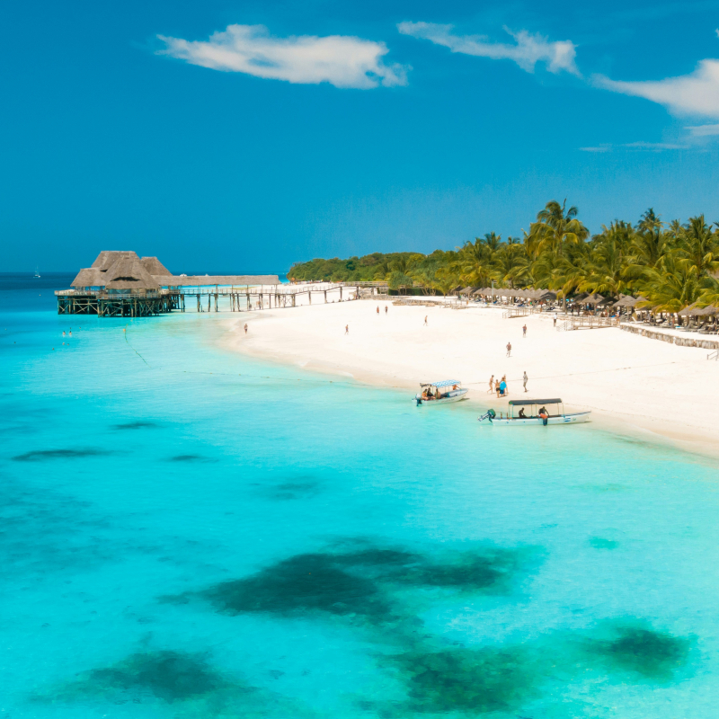 A high-angle view of a tropical beach resort featuring clear turquoise water, white sand, small boats, palm trees, and a wooden pier structure.