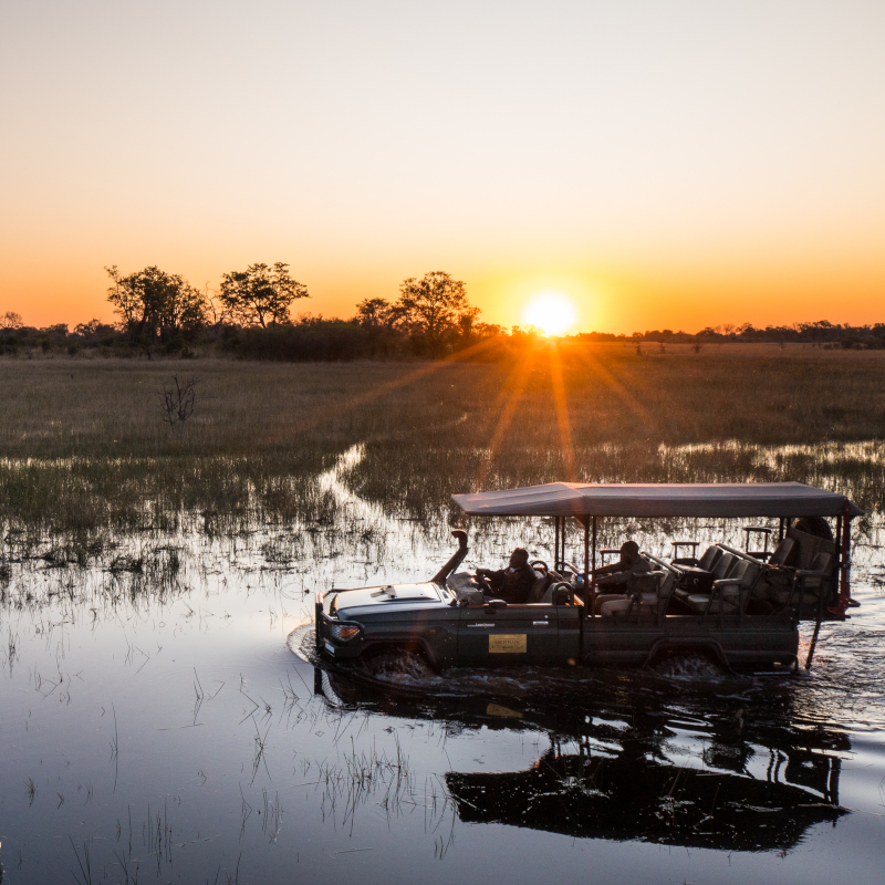 2022-GreatPlains-Okavango-Explorers-Camp-AHP-100 An open safari vehicle driving through shallow water at sunset.