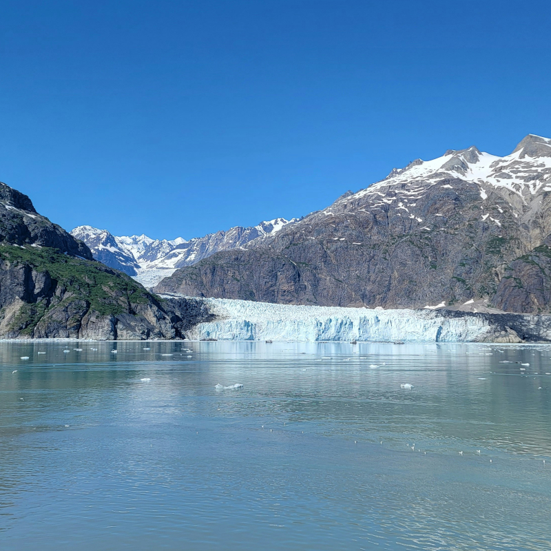 Alaska-Margerie-Glacier Wide view of a massive blue-ice tidewater glacier meeting calm ocean waters, flanked by rocky, snow-spotted mountains.
