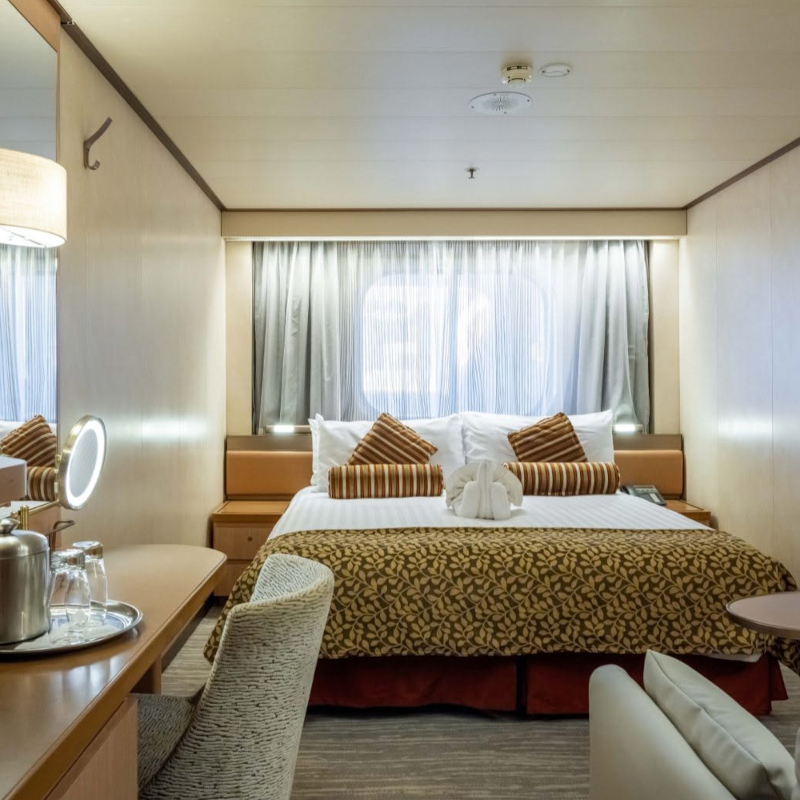 Ambassador-Renaissance-outside-cabin A cosy outside cabin bedroom on a cruise ship featuring a double bed, a vanity desk, and a window with light curtains
