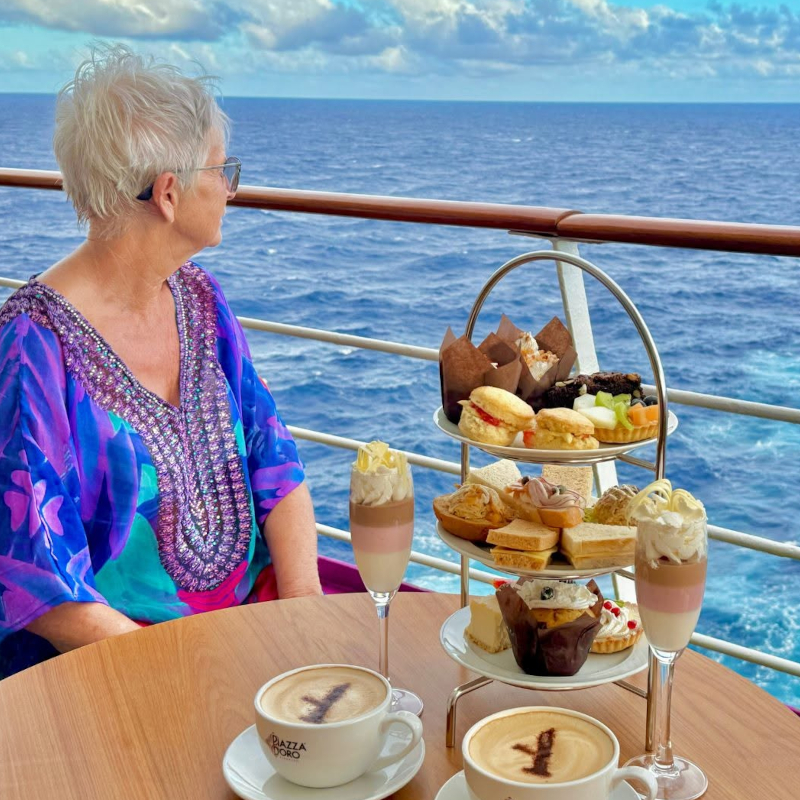 Ambassador-afternoon-tea A woman enjoying a multi-tiered afternoon tea stand with coffee on a cruise deck looking out at the ocean.