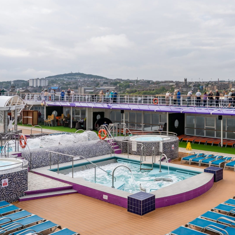 Ambassador-deck A cruise ship pool deck featuring bubbling hot tubs, blue lounge chairs, and passengers on an upper walkway overlooking a coastal city.