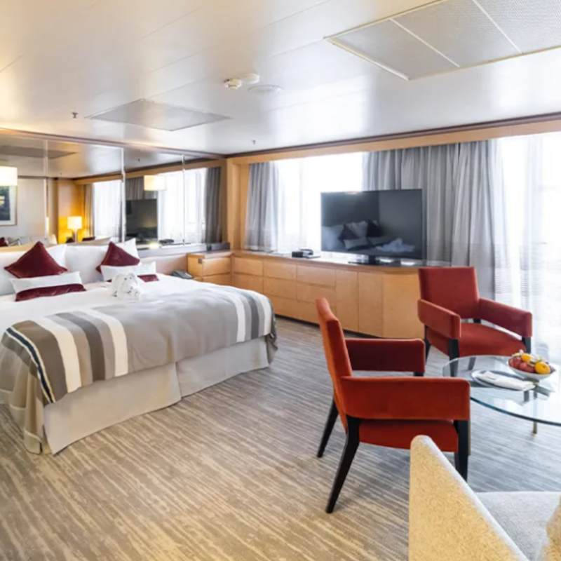 Ambassador-suite-Renaissance A spacious cruise ship suite featuring a large double bed, a seating area with two red armchairs, a flat-screen TV, and large windows.