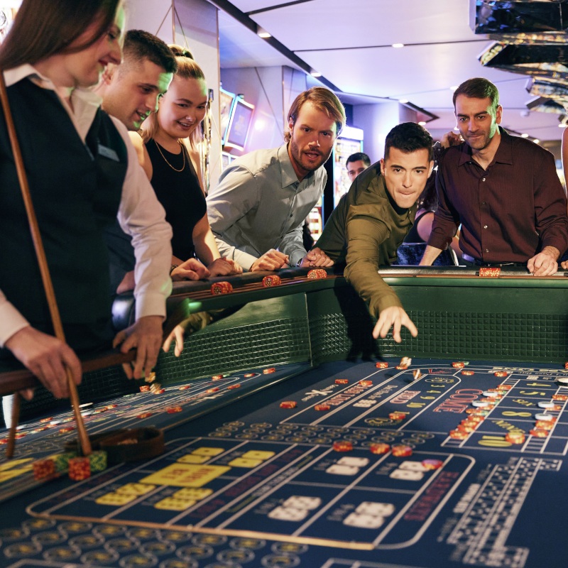 Celebrity-beyond-casino A group of people watching intently as a man rolls the dice on a vibrant blue craps table in a modern casino.