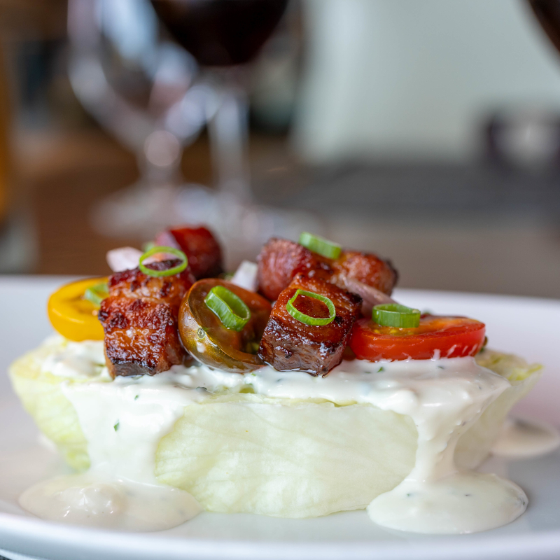 Celebrity-dining2 Close-up of a wedge salad topped with creamy blue cheese dressing, crispy pork belly cubes, sliced cherry tomatoes, and green onions.