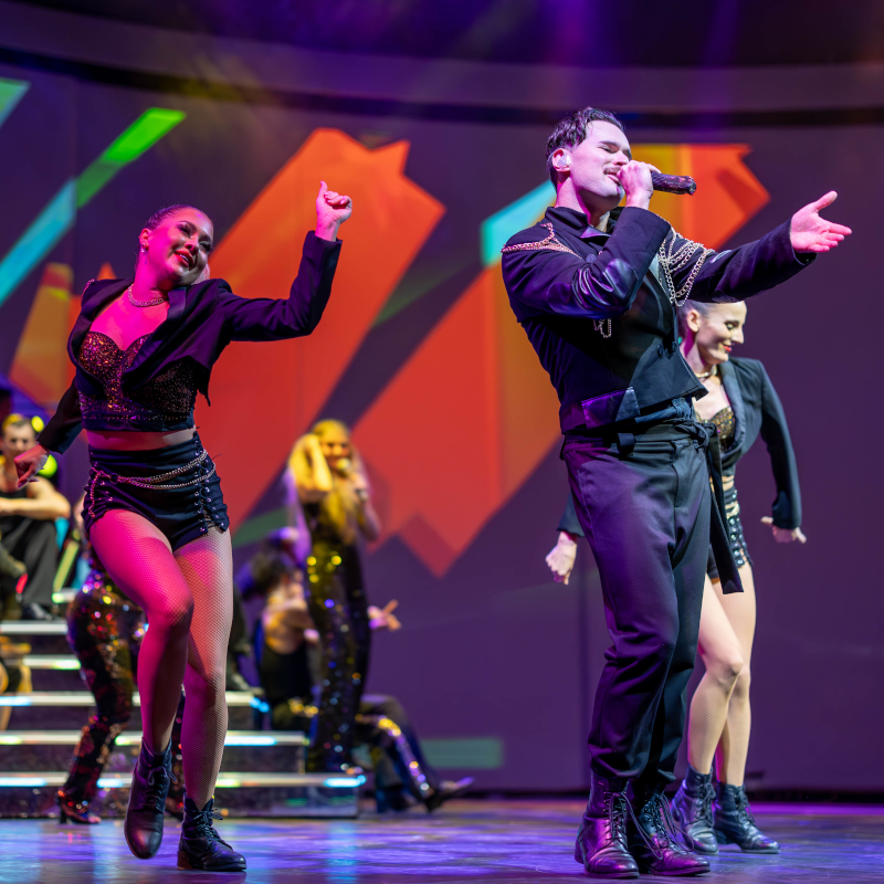 Celebrity-entertainment A male lead singer and energetic female dancers in stylish black outfits performing choreography on a brightly lit theatre stage.