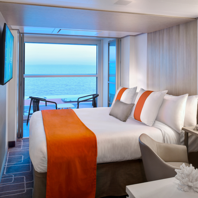 Celebrity-single-stateroom A modern cruise ship cabin featuring a bed with orange accent pillows and a runner, next to a large floor-to-ceiling glass window overlooking the ocean.