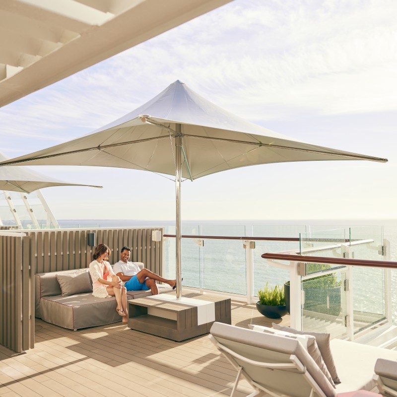 Celebrity-sundeck A couple relaxing on a premium wooden sun deck with comfortable outdoor lounge seating and a large white canopy umbrella overlooking the sea.