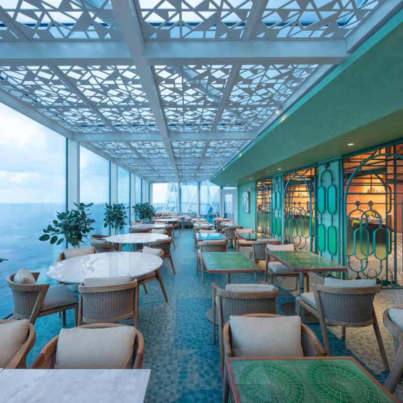 Celebrity-xcel-terrace An elegant cruise ship dining terrace with teal tiled floors, green and marble tables, woven chairs, and a geometric-patterned canopy ceiling overlooking the open ocean.