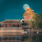 A traditional Chinese pavilion and corner tower at the edge of a calm moat with a large full moon in the night sky.