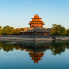 A corner tower of the Forbidden City with a multi-tiered roof perfectly reflecting on the smooth surface of a surrounding moat.