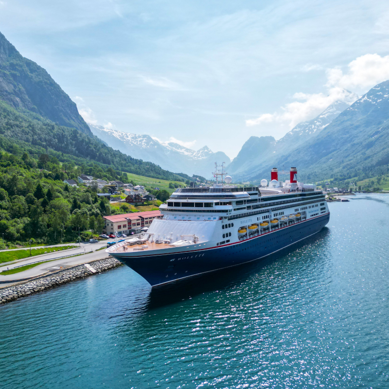 Fred-Olsen-Bolette-Norway The Bolette cruise ship gliding past a traditional red wooden house and green trees on the banks of a serene Norwegian fjord.